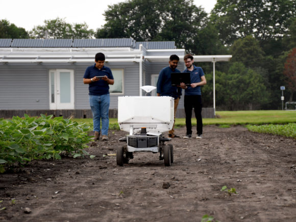 Photograph of three students holding a laptop and tablet controlling an white, calf-level high, autonomous farm robot as it moves through a soy bean field