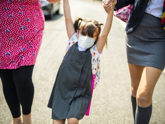 Photograph of a young girl wearing a mask swinging by her arms from two other individuals holder her up as they walk