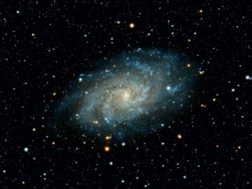 Photograph of the Triangulum Galaxy in outer space with hues and gradients of cyan, yellow, and white throughout with surrounding stars which glow in white and yellow