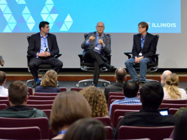 Photograph of five people in business attire sitting on stage during a symposium panel with the center person speaking into a microphone while all are sitting in front of a blue projection with the University of Illinois word mark