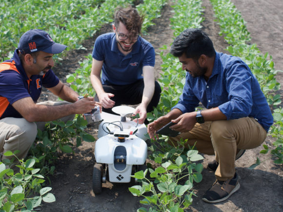Outdoor photograph of three individuals crouching in a soy bean field and talking about and working on a white autonomous farm robot that is calf-high