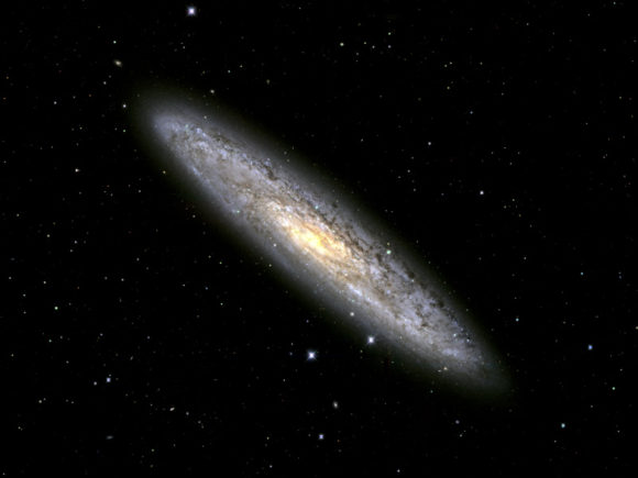Photograph of the Silver Dollar galaxy taken by the Dark Energy Survey Collaboration with the DECAM camera mounted on the Blanco Telescope at the Cerro Tololo Observatory in Chile. The galaxy is diagonal and flat with surrounding dust and stars seen in gradients of cyan, white, and yellow shining at the center
