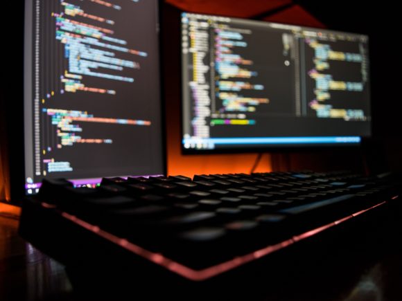 Black keyboard and monitors in soft focus with multicolored html code in front of an orange backlit background
