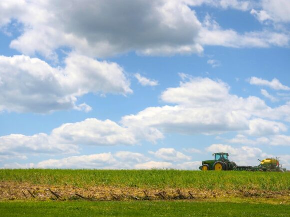 A farmer planting soybean plants in a field using a green and yellow John Deere tractor on a sunny day with big fluffy white clouds in a light blue sky.