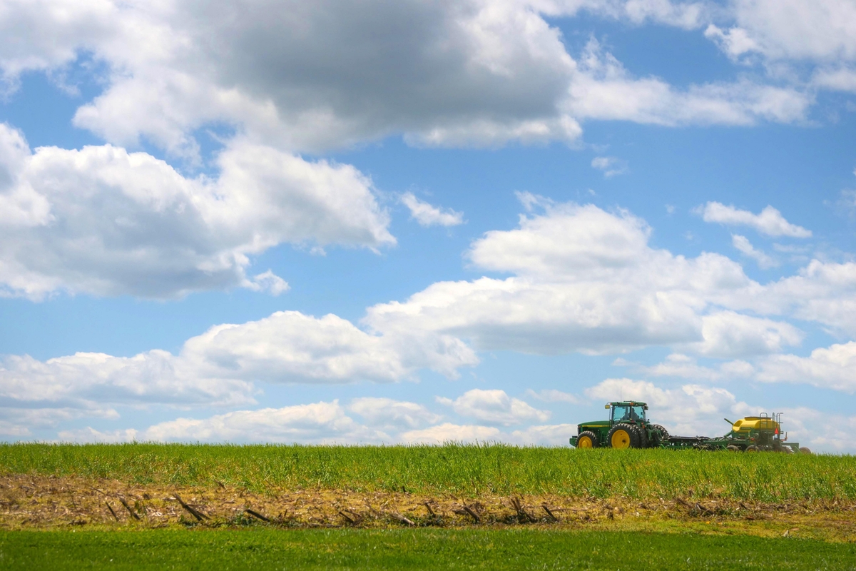 A farmer planting soybean plants in a field using a green and yellow John Deere tractor on a sunny day with big fluffy white clouds in a light blue sky.