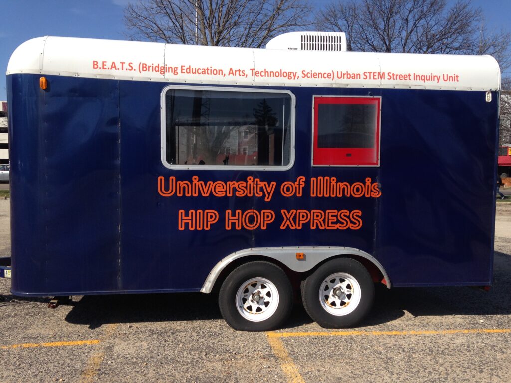 The second iteration of the Hip Hop Xpress, a blue and orange painted enclosed trailer with the following words painted on it. "B.E.A.T.S (Bridging Education, Arts, Technology, Science) Urban STEM Street Inquiry Unit" A large side window and an embedded TV face the viewer and the text "University of Illinois - Hip Hop Xpress