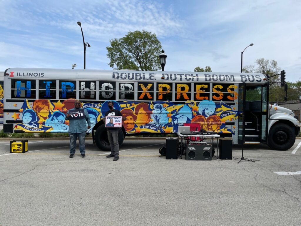 William Patterson standing outside in front of the stylized Hip Hop Xpress Double Dutch Boom Bus in a parking lot. holding "vote" signs