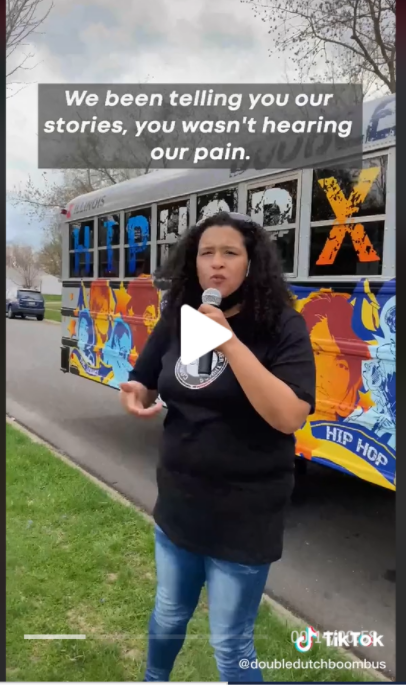  A screen capture of a TikTok music video. In foreground, a person of color raps "We been telling you our stories, you wasn't hearing our pain" while holding a microphone. In background, the Double Dutch Boom Bus.