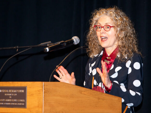 Photograph of Donna Cox speaking at a podium while wearing a white and black polka-dot blazer with red glasses; standing in front of a black back-drop curtain.
