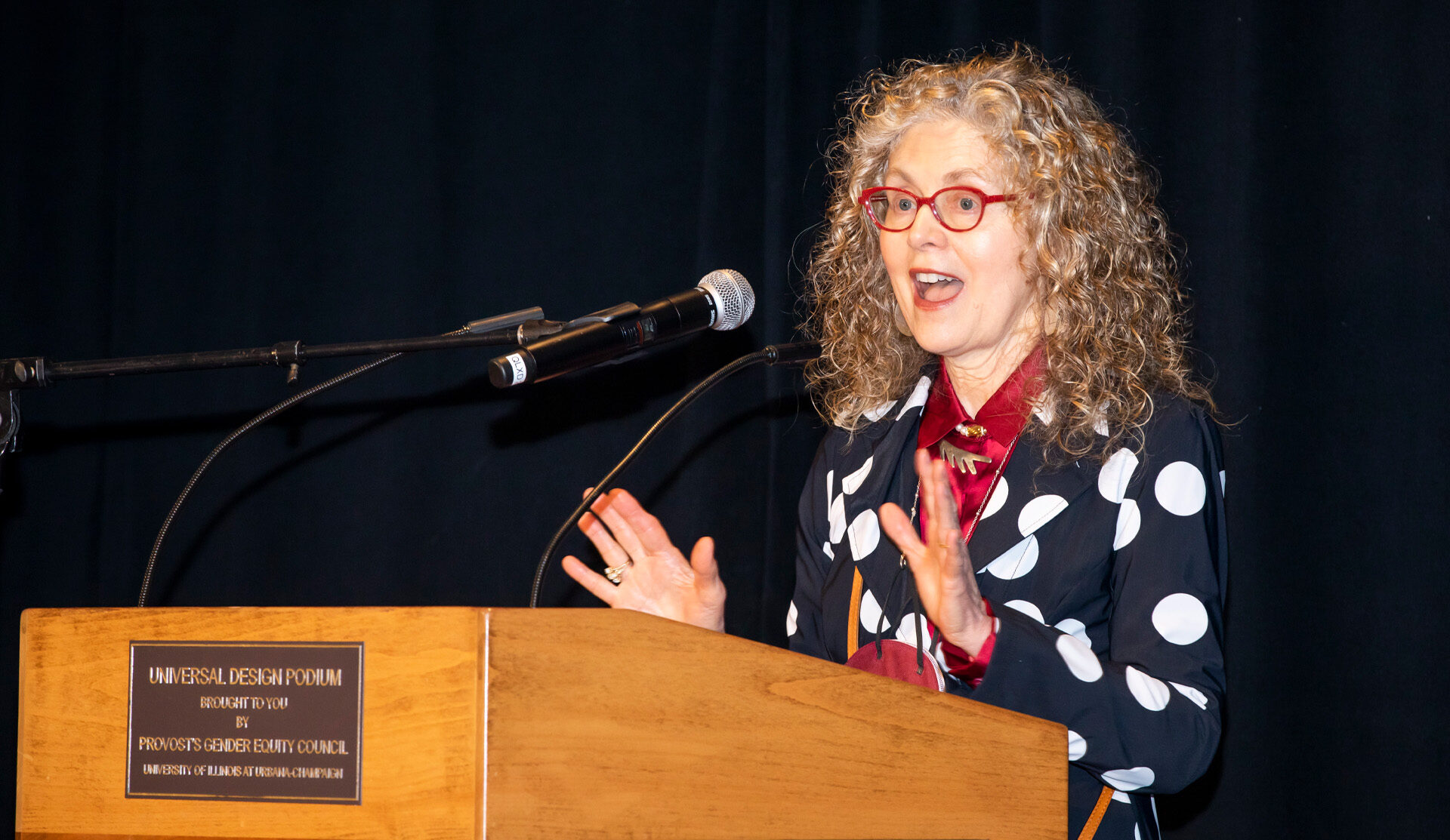 Photograph of Donna Cox speaking at a podium while wearing a white and black polka-dot blazer with red glasses; standing in front of a black back-drop curtain.
