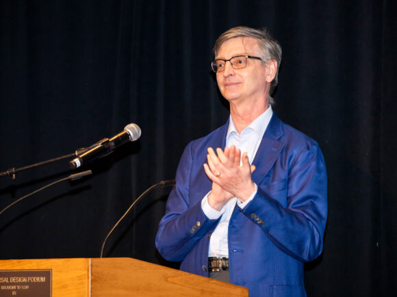Photograph of Bill Gropp in a blue jacket applauding and smiling at a podium; he is standing in front of a black back-drop.