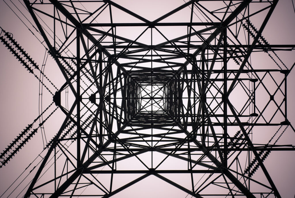 Abstract photograph taken from the ground looking directly up into electrical infrastructure. Dark lines of cables and metal contrast with the light sky.