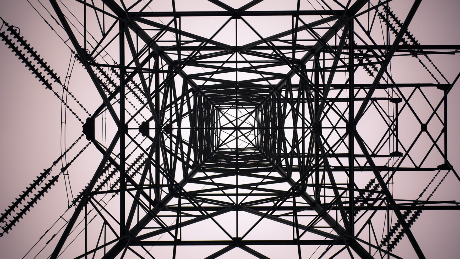 Abstract photograph taken from the ground looking directly up into electrical infrastructure. Dark lines of cables and metal contrast with the light sky.