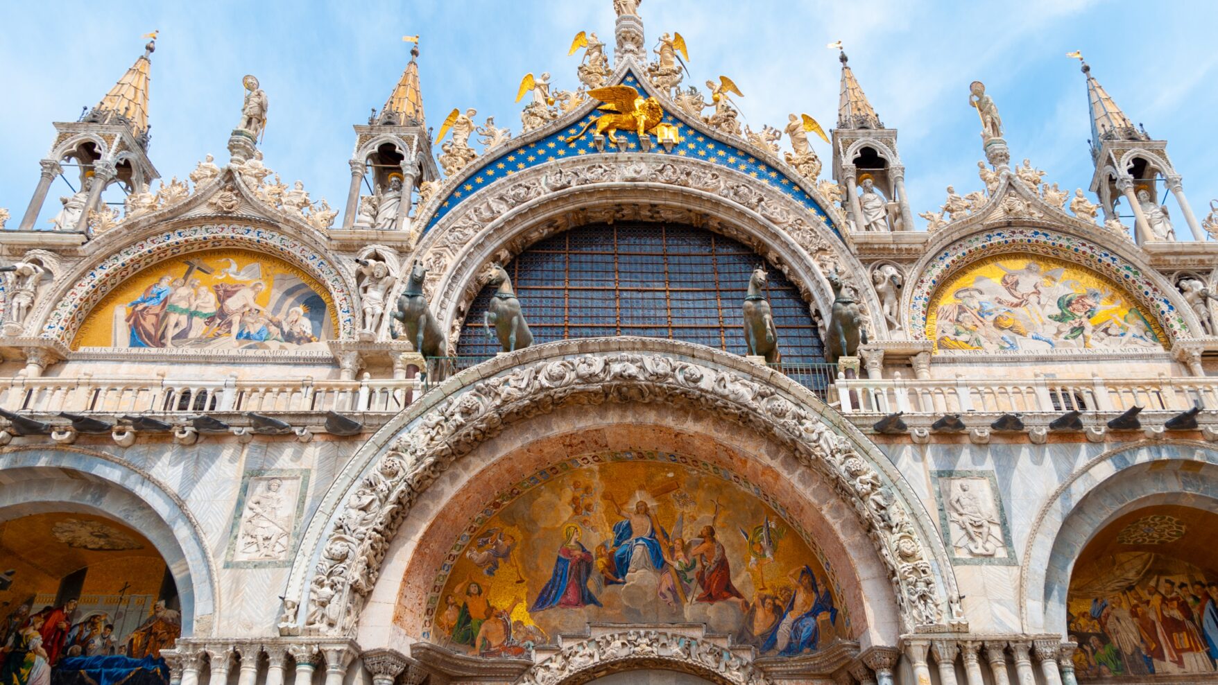 San Marco Basilica in Venice. Ornate statues, arches, and steeples with frescos and gold and blue decoration.