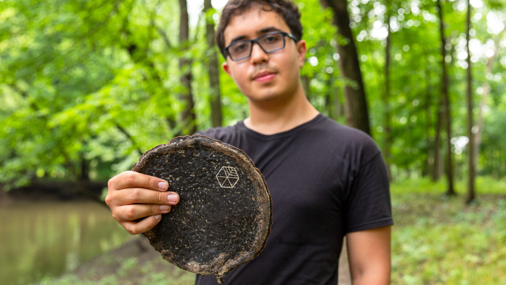 Gabe Tavas standing in a forest preserve surrounded by trees holding a rounded piece of Pyrus, the wood alternative.