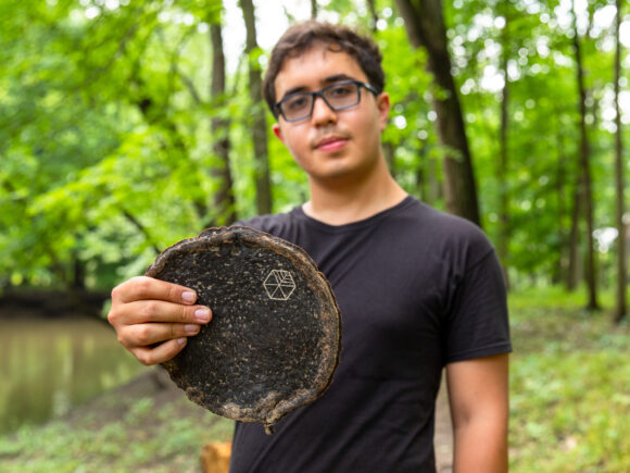 Gabe Tavas standing in a forest preserve surrounded by trees holding a rounded piece of Pyrus, the wood alternative.