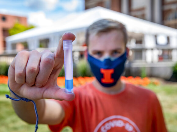 A UIUC student in an orange t-shirt and blue face mask holding a test tube with blue liquid.