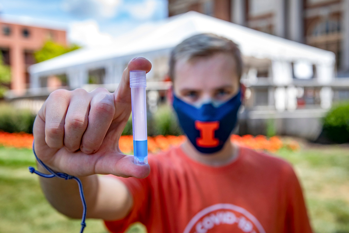 A UIUC student in an orange t-shirt and blue face mask holding a test tube with blue liquid.