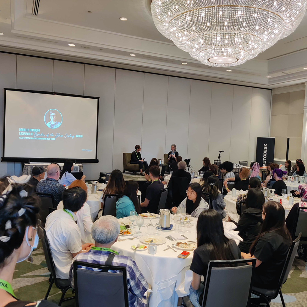 Multiple attendees seated at round tables listening to a presentation during the Women of SIGGRAPH Lunch Mixer.
