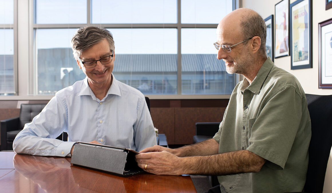 Director Bill Gropp and Chief Scientist Dan Katz sit at the end of a wooden conference table talking casually and smiling. Both are looking down and pointing at a tablet positioned between the two of them.