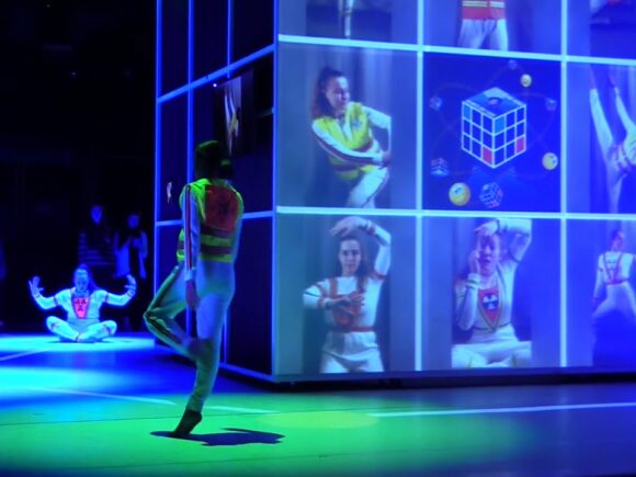 People dancing on a stage next to a 3x3 screened cube with images of posed dancers.