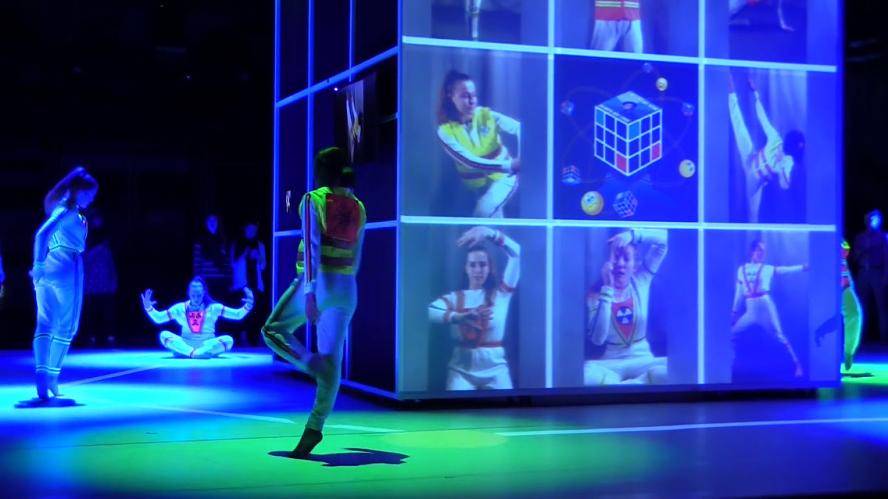 People dancing on a stage next to a 3x3 screened cube with images of posed dancers.
