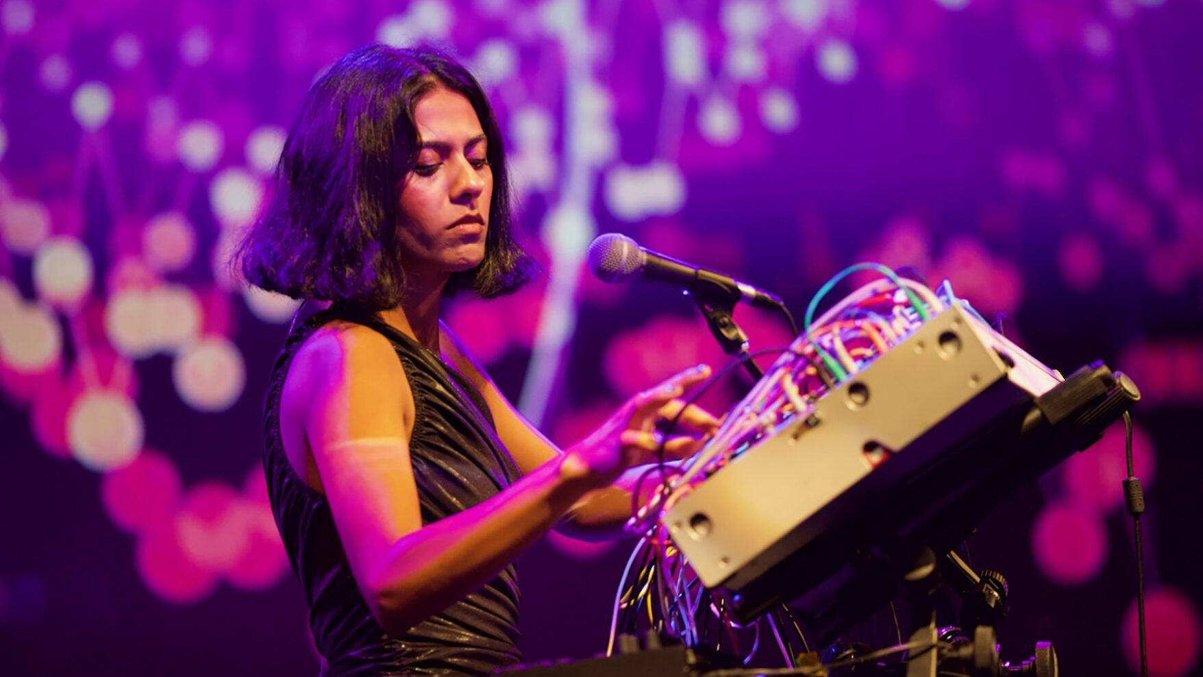 Arushi Jain performs at Krannert during Pygmalion 2023. She's standing in front of a large technical device with a lot of wires called a modular synthesizer. The screen behind her is full of purple tinted stars.