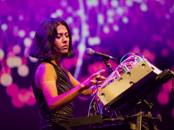 Arushi Jain performs at Krannert during Pygmalion 2023. She's standing in front of a large technical device with a lot of wires called a modular synthesizer. The screen behind her is full of purple tinted stars.