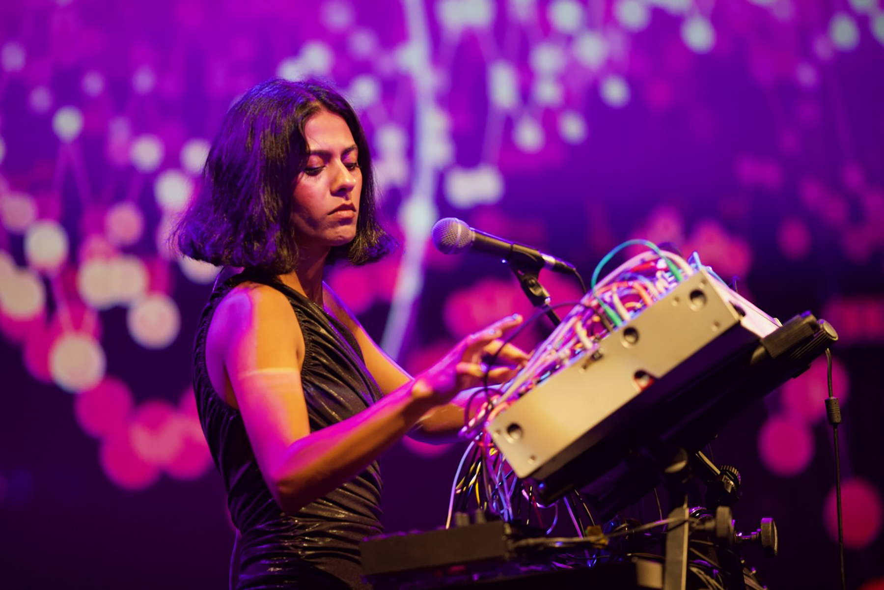 Arushi Jain performs at Krannert during Pygmalion 2023. She's standing in front of a large technical device with a lot of wires called a modular synthesizer. The screen behind her is full of purple tinted stars.