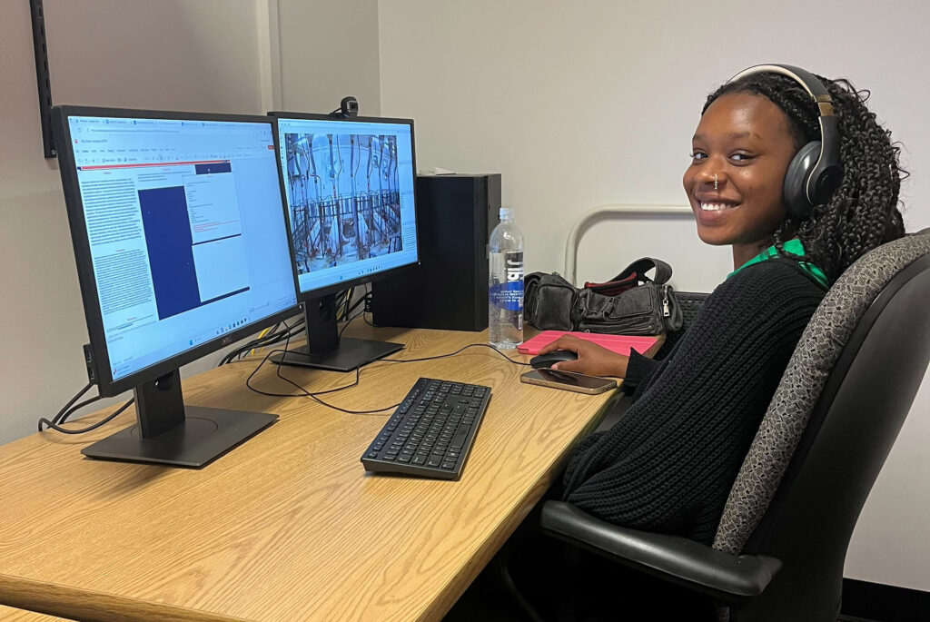 De'Janique Lambeth is sitting at a desk in front of a computer. She's turning to smile at the camera.